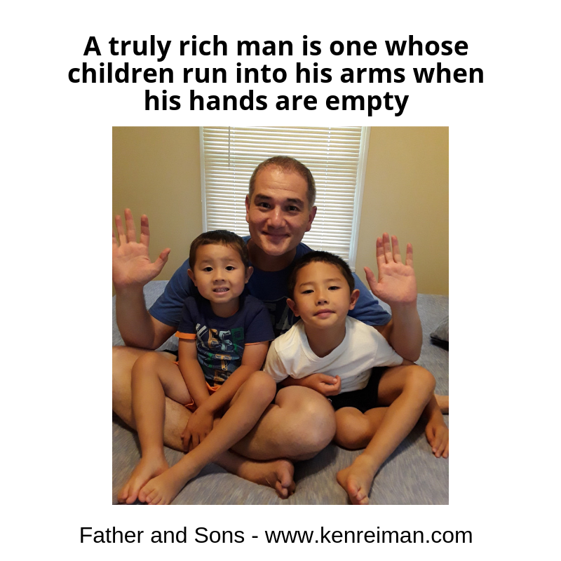 Father sitting on a bed with two young children, all smiling and raising their hands together.
